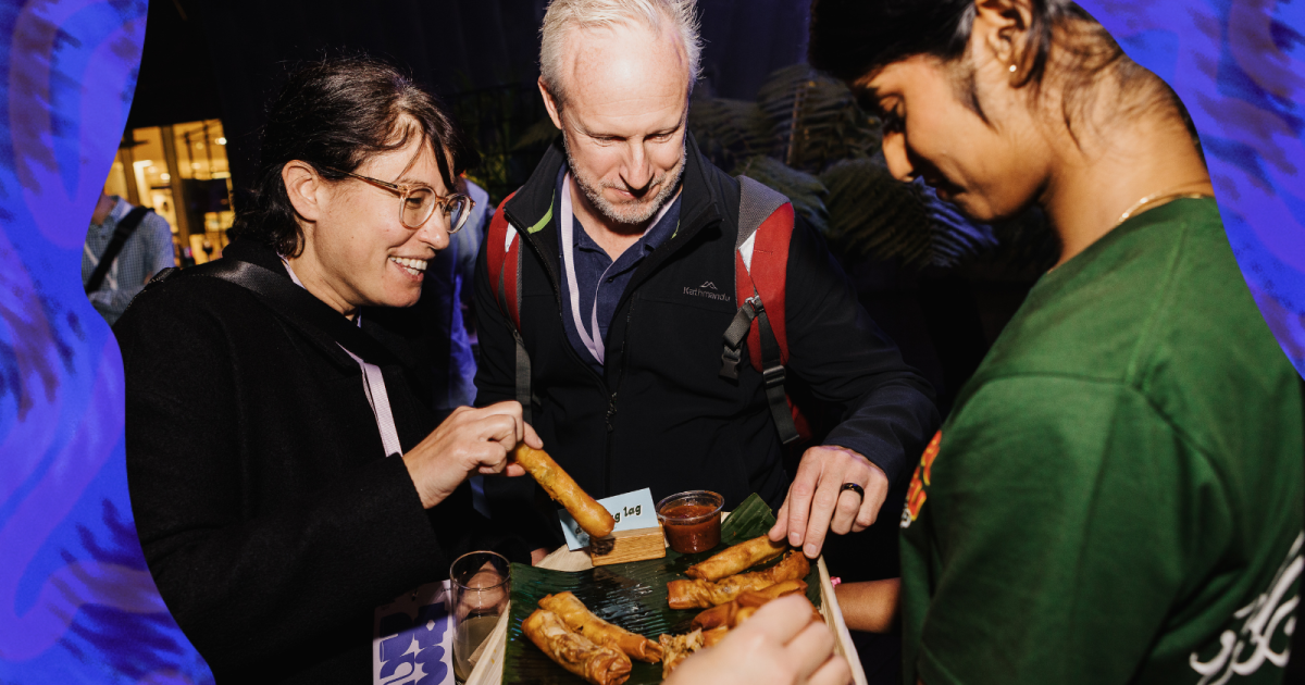 A group of three adults enjoying a social event. On the left, a woman with glasses and a black jacket is smiling as she holds a spring roll, poised for a dip in sauce. In the center, a man with short blond hair, wearing a red and gray jacket with a blue lanyard, is attentively looking at the food. On the right, another woman with her back partially turned to the camera is wearing a green shirt; she's also holding a spring roll near a sauce. They're standing around a table covered with a blue cloth and a bam