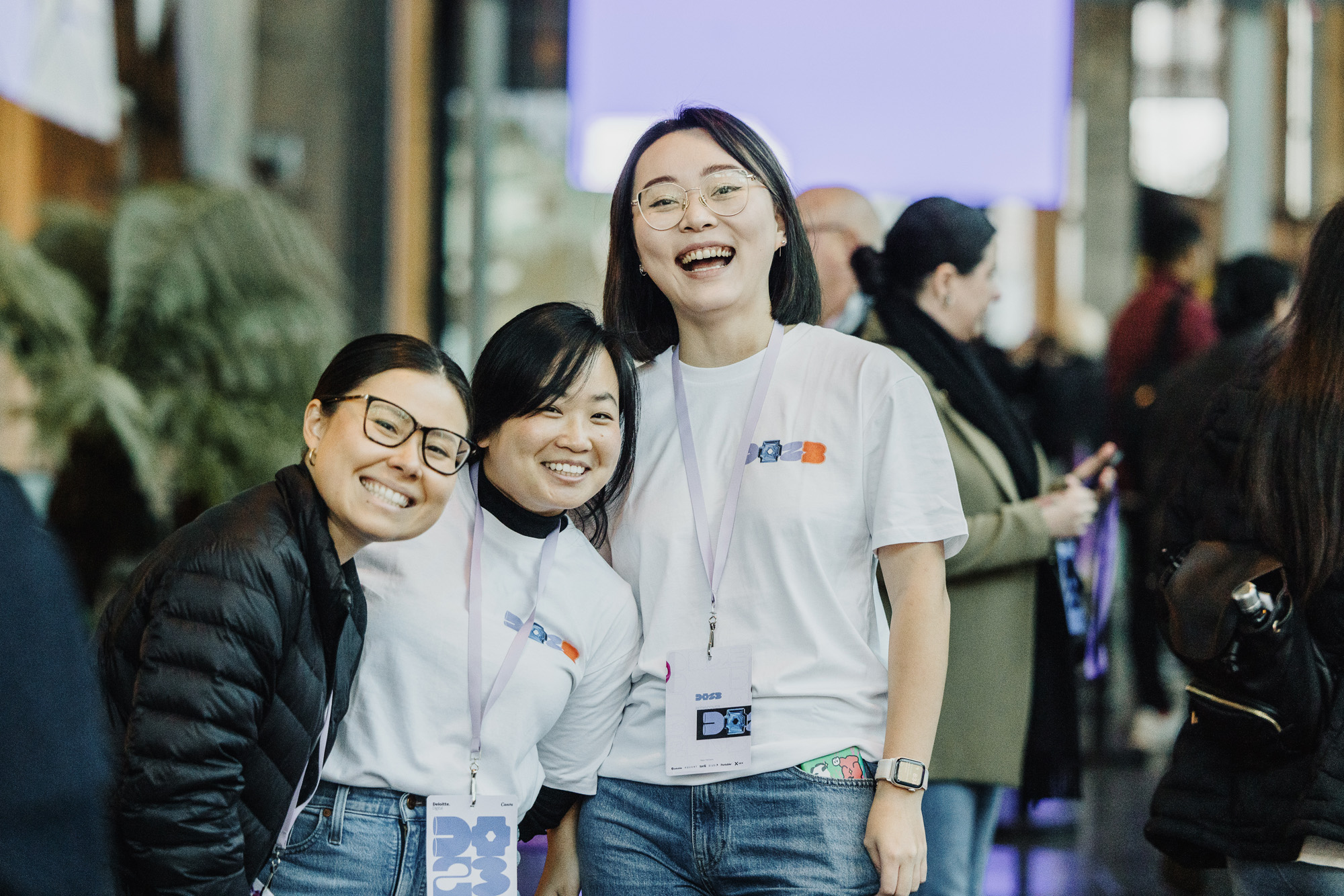 Three joyful volunteers in white t-shirts and lanyards, hugging and smiling at DO23